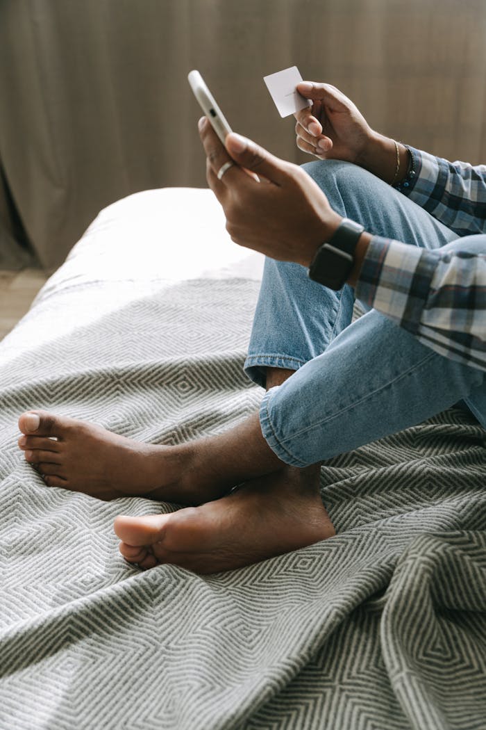 Person using smartphone for online shopping while sitting barefoot on bed.
