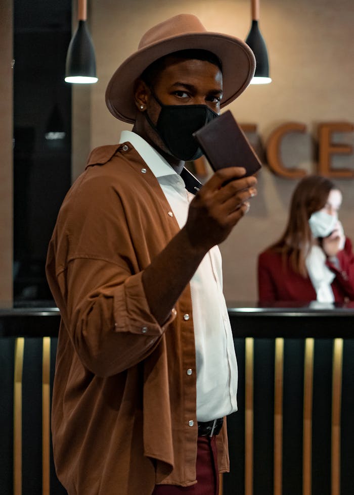 about-01 A man wearing a mask and hat holds a passport at a hotel reception, emphasizing travel safety.